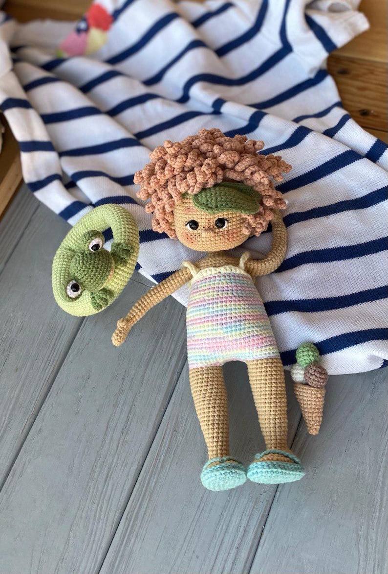 Pode incluir: Uma boneca de croch&ecirc; vestindo um mai&ocirc; listrado e segurando um cone de sorvete verde de croch&ecirc;. A boneca tem cabelo castanho encaracolado e usa sand&aacute;lias azuis. A boneca est&aacute; deitada em um cobertor listrado azul e branco.