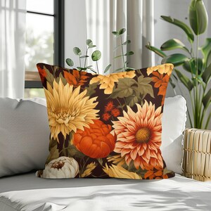 Op de afbeelding: Een vierkant sierkussen met een donkerbruine achtergrond en een dicht bloemenpatroon van grote gele, oranje en witte chrysanten en dahlia's, afgewisseld met groene bladeren en kleine rood-oranje stekelige bloemaccenten.