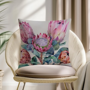Puede incluir: Un cojín decorativo con un diseño floral en acuarela. El cojín muestra flores de protea rosas con hojas y capullos verdes sobre un fondo neutro. El cojín está colocado sobre una silla color crema.