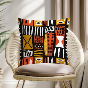 Peut inclure: Coussin carré avec un motif géométrique audacieux en noir, orange, blanc et or. Le design présente des formes et des lignes abstraites, créant un look moderne et artistique. Le coussin est posé sur une chaise beige.