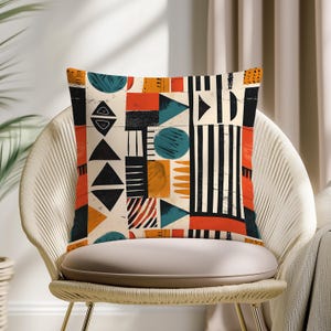May include: Square cushion with a geometric pattern in orange, black, teal, and cream. The design features triangles, circles, and stripes. The cushion is on a white chair.