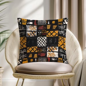 May include: Square throw pillow with a patchwork design in black, yellow, white, and red. The pillow is placed on a cream-colored chair with a woven back. The pillow's pattern features geometric shapes and lines.