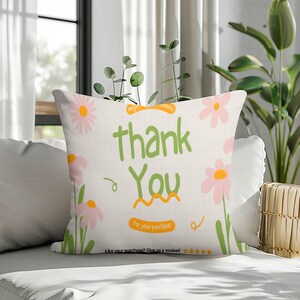 Op de afbeelding: Een vierkant sierkussen met een witte rasterachtergrond en illustraties van lichtroze madeliefjes met groene stelen. De tekst luidt thank You in het groen, met oranje accenten die For your purchase en Like your purchase? Give us a review vermelden.