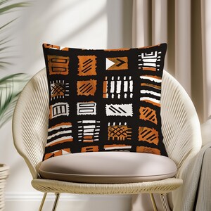 May include: Square throw pillow with a black background and a repeating pattern of orange, white, and brown geometric shapes. The pillow is placed on a beige chair with a woven back.