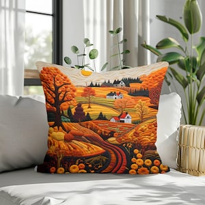 Op de afbeelding: Een vierkant sierkussen met een getextureerde illustratie in quiltstijl van een herfstlandschap. De scène toont glooiende heuvels, oranje en gele bomen, witte huizen met rode daken en een kronkelend pad omringd door pompoenen.