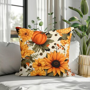 Op de afbeelding: Een vierkant sierkussen met een crèmekleurige achtergrond en een herhalend patroon van feloranje pompoenen, grote gele zonnebloemen, kleinere gouden bloesems, donkergroen gebladerte en kleine bruine bessen over het hele ontwerp.