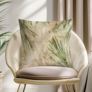 May include: Square cushion with a nature-inspired design. The cushion features a watercolour-style print of green pine needles and other foliage against a beige and brown background. The cushion is placed on a woven chair.