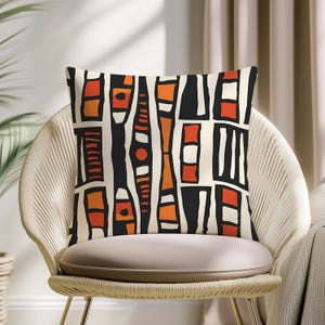 May include: Square decorative pillow with an abstract geometric pattern in black, orange, and white. The pillow is on a beige chair with a woven back. The design features vertical and horizontal shapes.