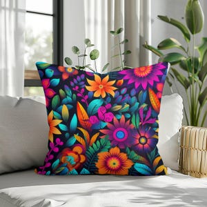 Op de afbeelding: Vierkant sierkussen met een levendig bloemenpatroon. Het ontwerp toont kleurrijke bloemen in tinten oranje, roze, paars en teal tegen een donkere achtergrond. Het kussen ligt op een witte bank, met een plant op de achtergrond.