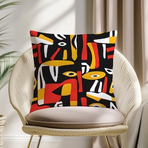 May include: Square throw pillow with an abstract design. The pillow features a black background with a pattern of red, yellow, and white shapes. The pillow is placed on a woven chair.