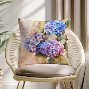 Könnte beinhalten: Quadratisches Zierkissen mit einem Aquarellgemälde von blauen und lila Hortensienblüten mit grünen Blättern. Das Kissen hat einen beigen Hintergrund und liegt auf einem geflochtenen Stuhl.