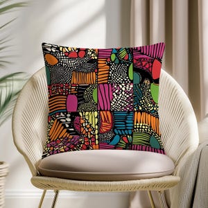 May include: A square throw pillow with a vibrant, geometric pattern in a patchwork design. The design features a mix of colors including orange, pink, green, and black. The pillow is placed on a white chair.