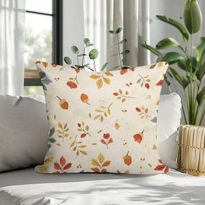 May include: A square throw pillow featuring a cream-coloured background with a repeating pattern of autumn-themed illustrations, including orange acorns, yellow and red leaves, and small berries scattered across the fabric surface.