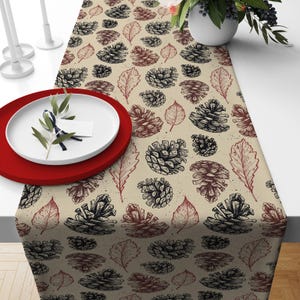 May include: A beige table runner with a repeating pattern of black and brown pine cones and leaves. The runner is on a white table, with a red plate and white plate with a sprig of greenery.