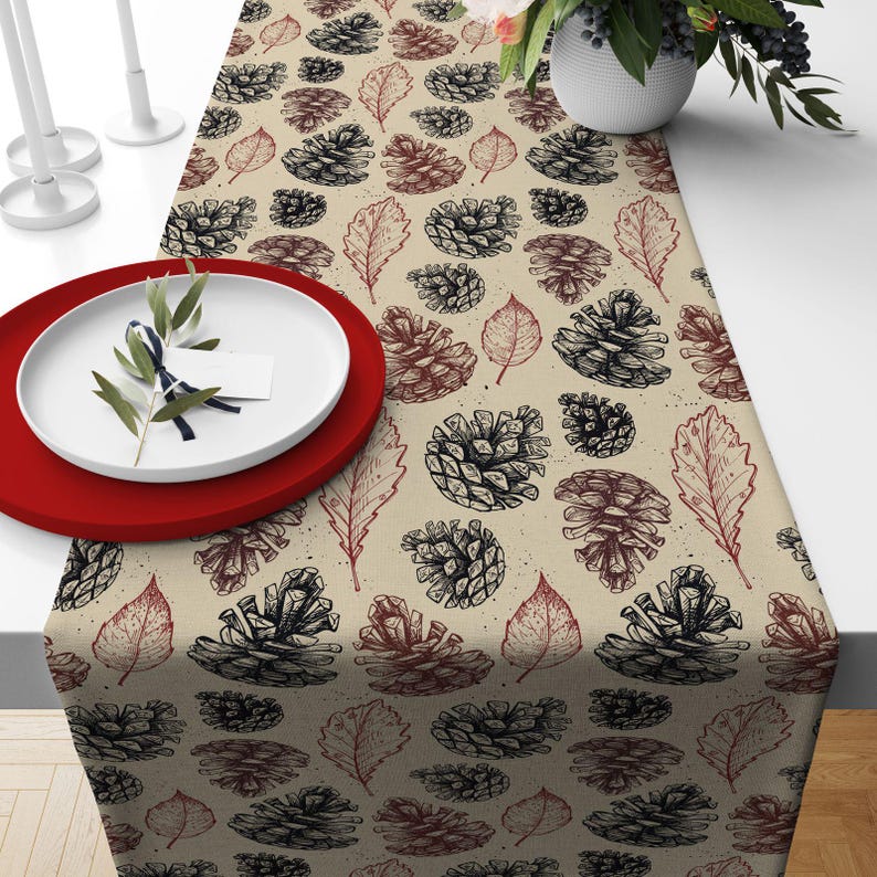 May include: A beige table runner with a repeating pattern of black and brown pine cones and leaves. The runner is on a white table, with a red plate and white plate with a sprig of greenery.