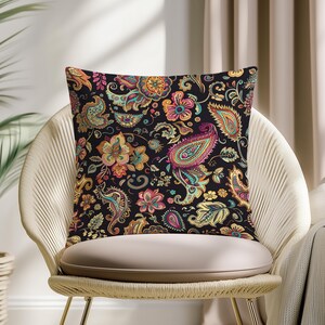 Op de afbeelding: Vierkant sierkussen met een zwarte achtergrond en een kleurrijk paisley bloemenpatroon. Het ontwerp toont bloemen en paisley vormen in tinten oranje, roze, blauwgroen en geel. Het kussen ligt op een geweven stoel.