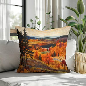 May include: A square throw pillow featuring a landscape print of a forest during autumn. The scene displays tall dark pine trees in the foreground, rolling hills with vibrant orange and yellow foliage, and distant blue mountains under a pale sky.