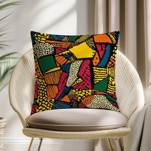 May include: A square throw pillow with a vibrant, abstract patchwork design. The pillow features a mix of geometric shapes in various colors, including yellow, red, green, orange, and black. The pillow is placed on a cream-colored chair.