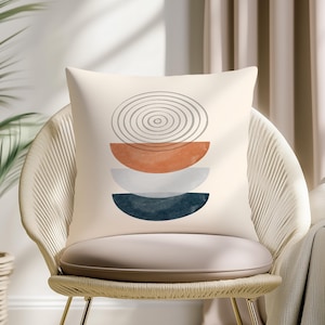 May include: Cream-coloured cushion with an abstract design. The design features semi-circles in orange, grey, blue, and white, topped with a grey spiral. The cushion is on a woven chair.