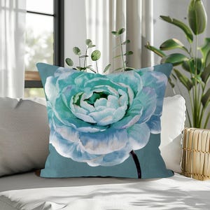 May include: A square throw pillow featuring a large, watercolor-style peony in shades of teal and light blue. The floral design is set against a teal background. The pillow is placed on a white sofa.