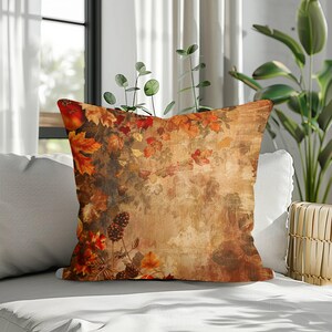 May include: A square throw pillow featuring a printed design of autumn foliage, including orange, red, and yellow maple leaves, along with dark brown pinecones. The background has a textured, weathered, and distressed tan and beige appearance.