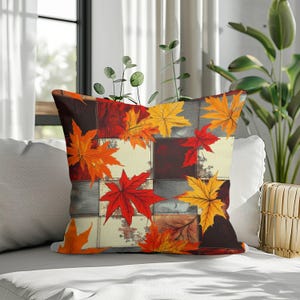 May include: A square throw cushion featuring a patchwork design of cream, dark brown, and grey squares. The surface is decorated with vibrant autumn maple leaves in shades of bright orange, deep red, and golden yellow scattered across the pattern.
