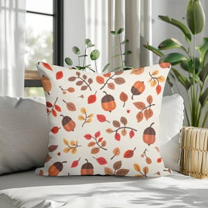 May include: A square throw cushion featuring a cream-coloured background with a repeating pattern of orange acorns, brown twigs, and various autumn-coloured leaves in shades of red, orange, yellow, and brown scattered across the entire surface.
