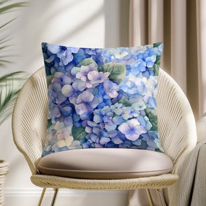 Può includere: Cuscino quadrato con un design ad acquerello di fiori di ortensia blu e viola con foglie verdi. Il cuscino è su una sedia beige con schienale intrecciato e un cuscino colorato chiaro. Il tema generale è floreale e l'arredamento della casa.