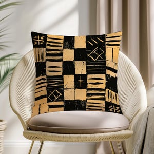 May include: Square cushion with a black and gold geometric pattern. The design features a patchwork of squares with various abstract shapes and lines. The cushion is placed on a woven chair.