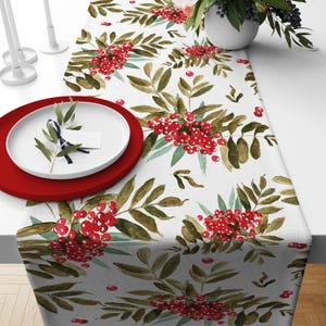 May include: A white table runner with a watercolor design of red berries and green leaves. A red plate and a white plate with a sprig of greenery are on the table. The runner is decorated with a repeating pattern.