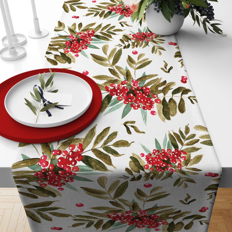 May include: A white table runner with a watercolor design of red berries and green leaves. A red plate and a white plate with a sprig of greenery are on the table. The runner is decorated with a repeating pattern.