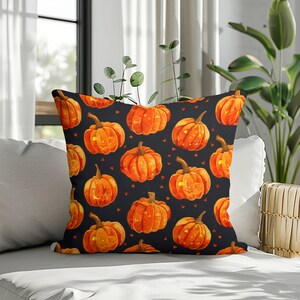 Op de afbeelding: Een vierkant sierkussen met een herhalend patroon van oranje pompoenen op een zwarte achtergrond. De pompoenen hebben uitgesneden gezichten met gloeiende gele ogen en monden, afgewisseld met kleine oranje driehoekige vormen.