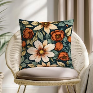 Puede incluir: Cojín decorativo con estampado floral. El cojín presenta grandes flores blancas y naranjas, capullos naranjas y hojas verdes sobre un fondo azul oscuro. Se coloca sobre una silla beige.