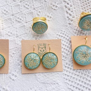 May include: Set of three gold rings and two pairs of earrings with delicate pressed flower designs in a light blue resin. The rings and earrings feature a gold metal setting.
