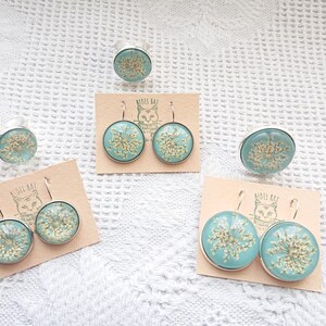 May include: Three sets of earrings and a ring with a light blue background and gold accents. The earrings are silver with a light blue background and gold accents. The ring is silver with a light blue background and gold accents. The earrings and ring are on a white background.