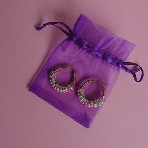 Pode incluir: Um par de brincos de argola dourados com contas coloridas, em uma bolsa de cord&atilde;o roxo.
