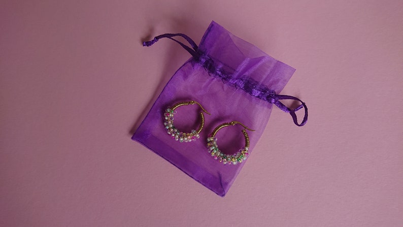 Pode incluir: Um par de brincos de argola dourados com contas coloridas, em uma bolsa de cord&atilde;o roxo.