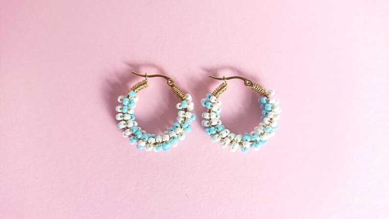 May include: A pair of gold hoop earrings with white and light blue beads. The beads are arranged in a spiral pattern.