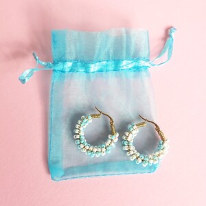 May include: A pair of hoop earrings made with white and light blue beads, displayed in a light blue organza bag.