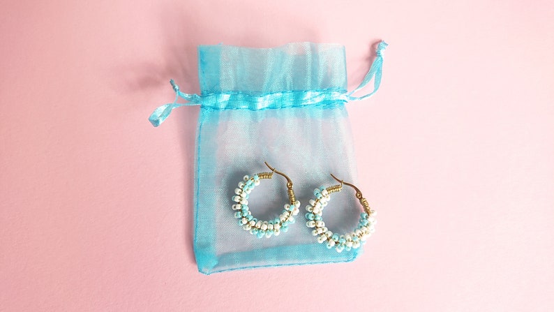 May include: A pair of hoop earrings made with white and light blue beads, displayed in a light blue organza bag.