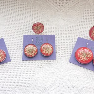 May include: Three sets of earrings and rings featuring pressed flowers encased in red resin. Each set includes a pair of earrings and a ring.