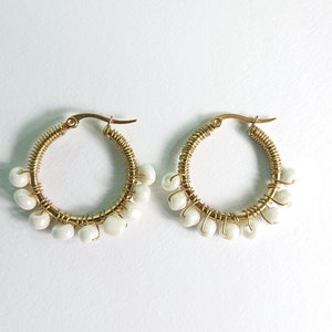 Pode incluir: Um par de brincos de argola dourados. Cada brinco é adornado com contas brancas redondas e enrolado com arame dourado. Os brincos são exibidos em um fundo branco.