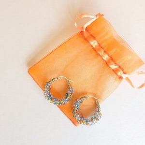 May include: A pair of gold hoop earrings with colourful beads. The earrings are in a small orange organza bag.