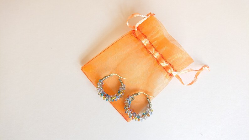 May include: A pair of gold hoop earrings with colourful beads. The earrings are in a small orange organza bag.