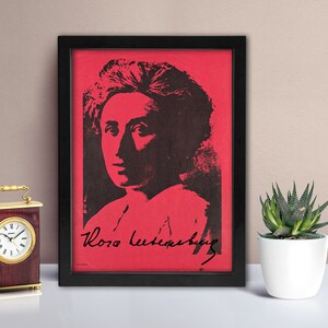 May include: A framed rectangular print featuring a high-contrast black portrait of Rosa Luxemburg set against a vibrant red background. The print includes the signature of Rosa Luxemburg in black script at the bottom, all encased in a black frame.