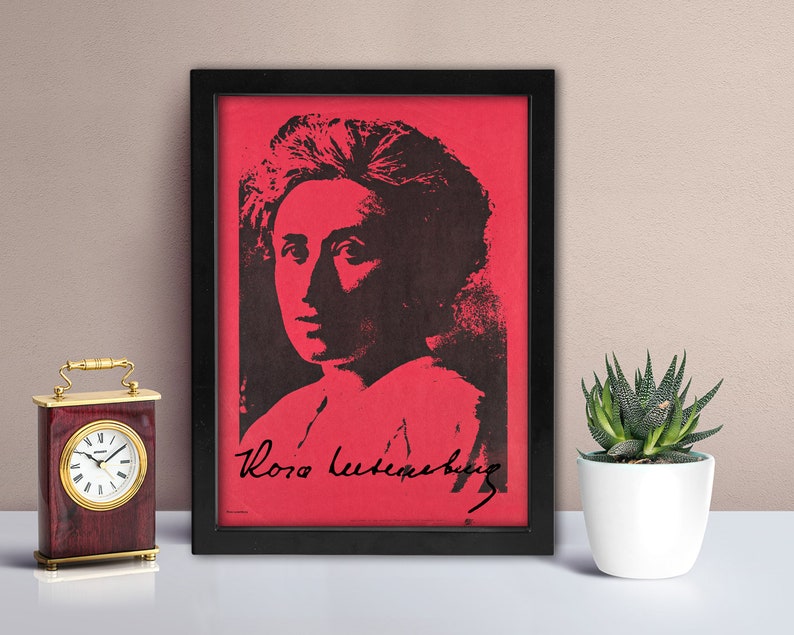 May include: A framed rectangular print featuring a high-contrast black portrait of Rosa Luxemburg set against a vibrant red background. The print includes the signature of Rosa Luxemburg in black script at the bottom, all encased in a black frame.