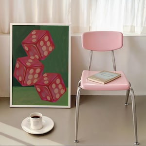 May include: A framed art print featuring three red dice with white dots against a green background. A pink chair with a book on the seat and a cup of coffee on a white plate complete the scene.