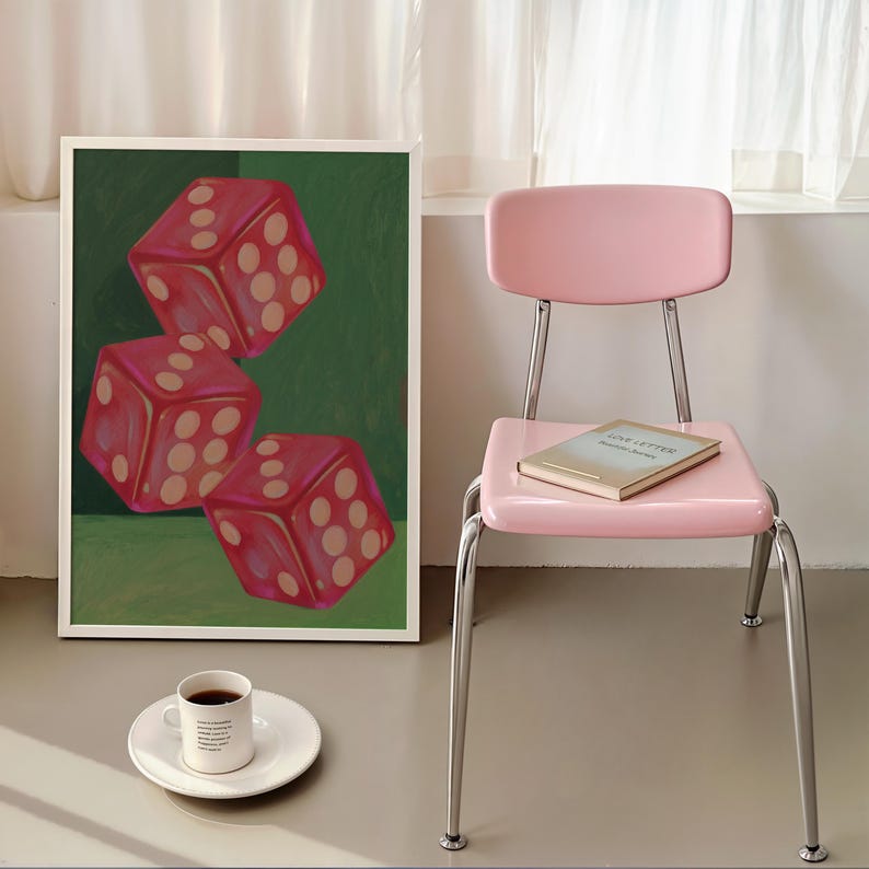 May include: A framed art print featuring three red dice with white dots against a green background. A pink chair with a book on the seat and a cup of coffee on a white plate complete the scene.