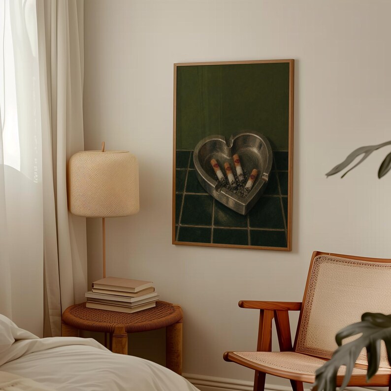 May include: A framed wall art print featuring a heart-shaped metal ashtray resting on a dark green tiled surface. Inside the ashtray are four lit cigarettes with visible ash and embers, set against a solid dark green background.