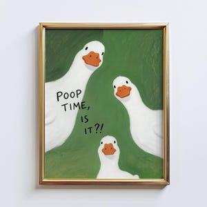 May include: A framed art print featuring three white geese with orange beaks against a green background. The text "POOP TIME, IS IT?!" is written on the print. The frame is gold.
